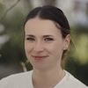 Eine Frau mit braunen Haaren und blauen Augen lächelt in die Kamera. Sie trägt ein weißes Oberteil und hat eine Halskette mit einem Anhänger. Der Hintergrund ist unscharf.