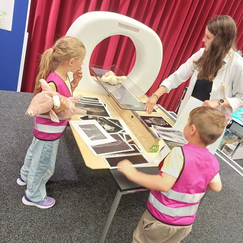 Eine Frau und zwei Kinder betrachten Röntgenaufnahmen auf einem Tisch, mit einem Spielzeug und einem Scanner im Hintergrund.