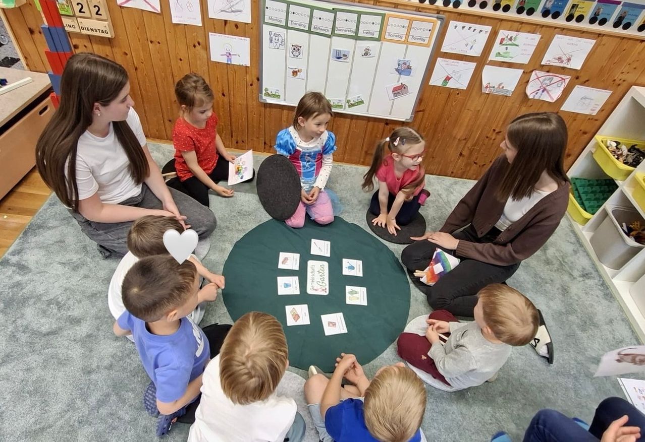 Kinder sitzen um einen grünen Kreis herum und sind mit Karten beschäftigt. Eine Frau ist bei ihnen. Der Raum hat eine Holzwand mit einer Tafel, die verschiedene Zeichnungen und Texte zeigt.