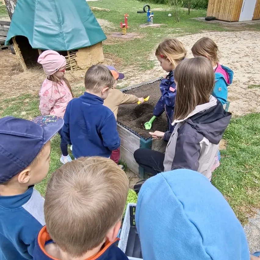 Mehrere Kinder spielen in einem Sandkasten in einem Spielplatz. Sie tragen Winterkleidung. Einige Kinder stehen, während andere sitzen. Es gibt einige Spielzeuge im Sand.