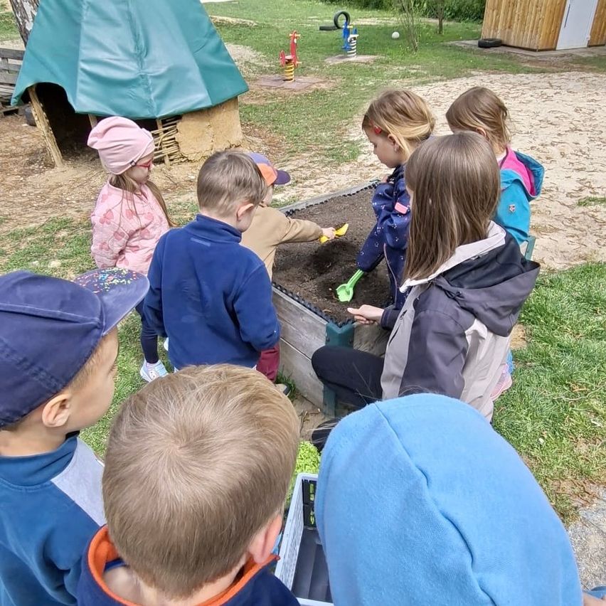 Mehrere Kinder spielen in einem Sandkasten in einem Spielplatz. Sie tragen Winterkleidung. Einige Kinder stehen, während andere sitzen. Es gibt einige Spielzeuge im Sand.