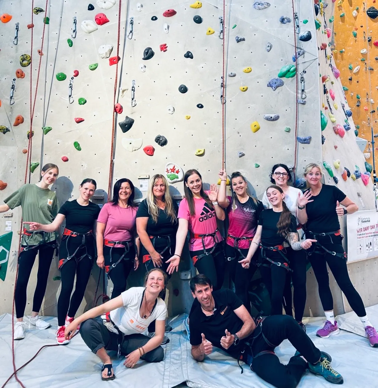 Eine Gruppe von Kletterern posiert für ein Foto vor einer Kletterwand mit verschiedenen farbigen Griffen und Haltegriffen. Einige sitzen, andere stehen.