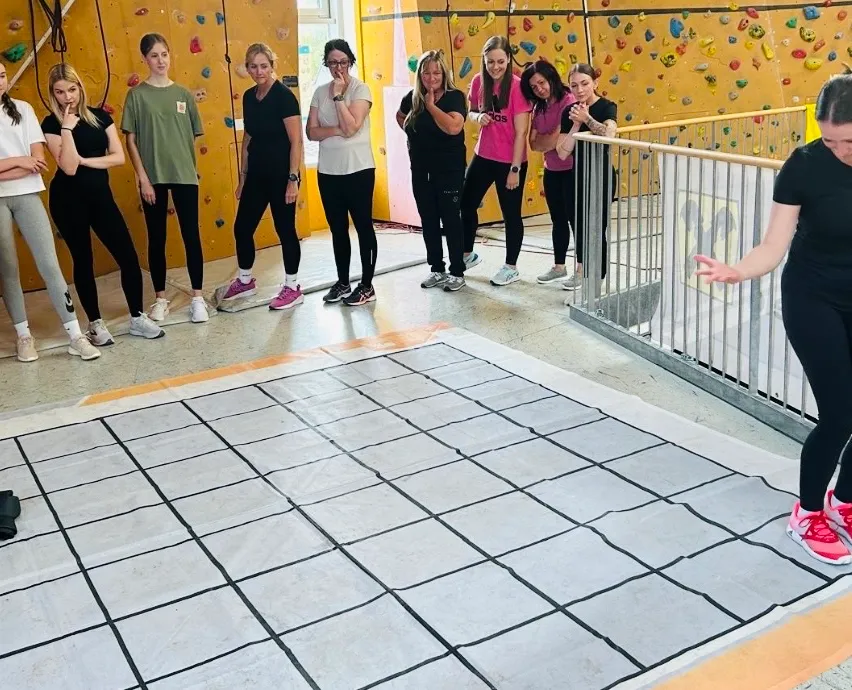 Mehrere Frauen stehen in der Nähe einer Kletterwand, tragen Sportkleidung und Turnschuhe. Eine Frau hält ihre Hand an den Mund, und eine andere lehnt sich gegen die Wand. Ein gefliester Boden befindet sich vor ihnen.