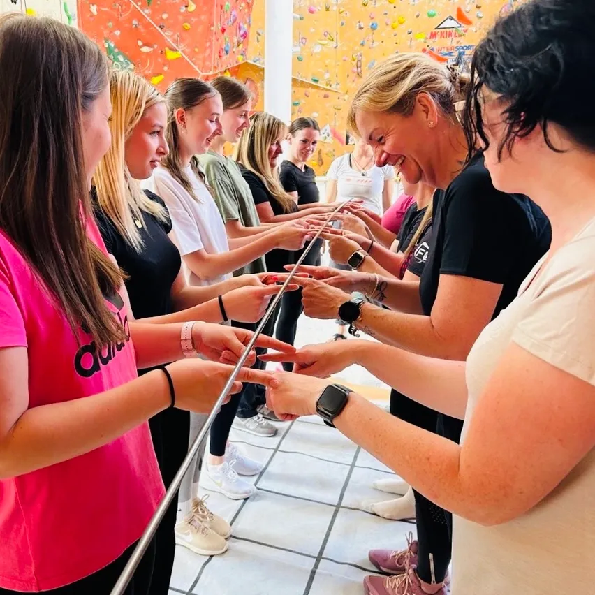 Eine Gruppe von Frauen hält sich an den Händen in einer Reihe in einem Fitnessstudio. Sie lächeln und schauen nach vorne. Sie tragen alle Turnschuhe und einige haben Armbänder.