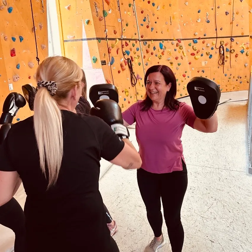 Frauen boxen mit Handschuhen in einem Fitnessstudio. Eine Frau in Pink lächelt und hält Boxhandschuhe.