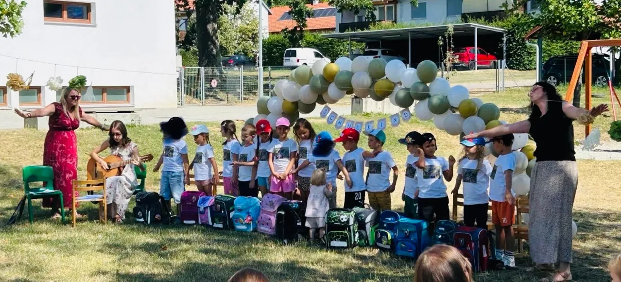 Eine Gruppe von Kindern in gleichen weißen T-Shirts steht in einer Reihe mit ihren Rucksäcken vor einem farbenfrohen Luftballonbogen.