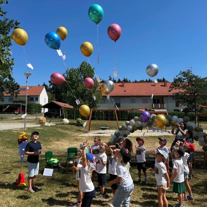 Eine Gruppe von Kindern auf einem Rasenfeld mit bunten Luftballons, die in der Luft schweben. Sie tragen weiße Hemden und Mützen. Hinter ihnen steht ein Gebäude mit einem roten Dach und einem Spielplatz.