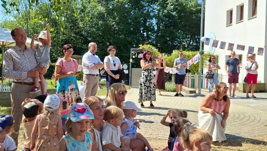 Eine Gruppe von Menschen, darunter Erwachsene und Kinder, versammelt sich im Freien. Einige stehen, andere sitzen auf dem Boden. Sie alle lächeln und scheinen die Veranstaltung zu genießen. Im Hintergrund befinden sich Bäume und ein Gebäude.