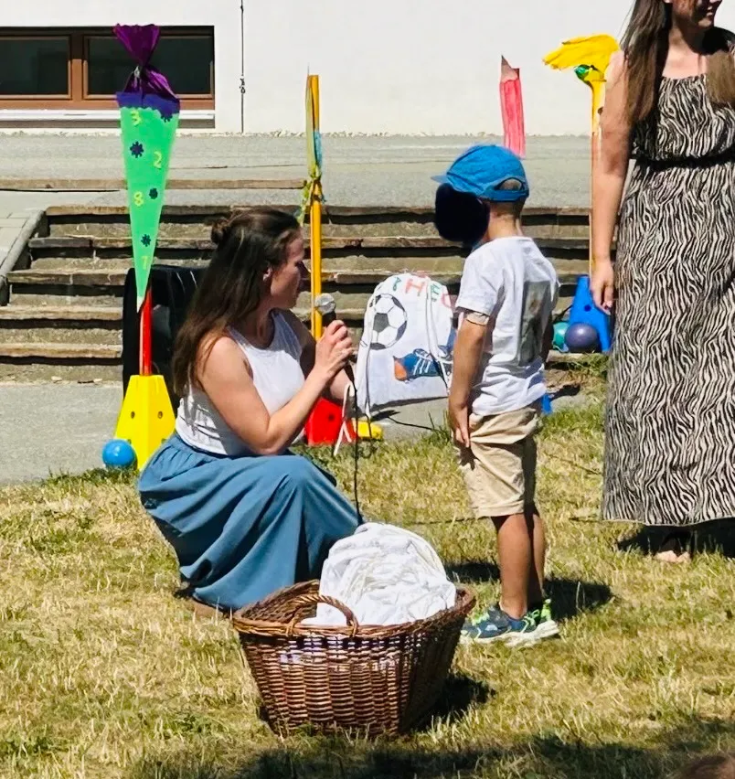 Eine Frau hält ein Mikrofon und spricht zu einem jungen Jungen in einem blauen Hut. Sie sind in einem Grasbereich mit verschiedenen bunten Objekten und einem Gebäude im Hintergrund.
