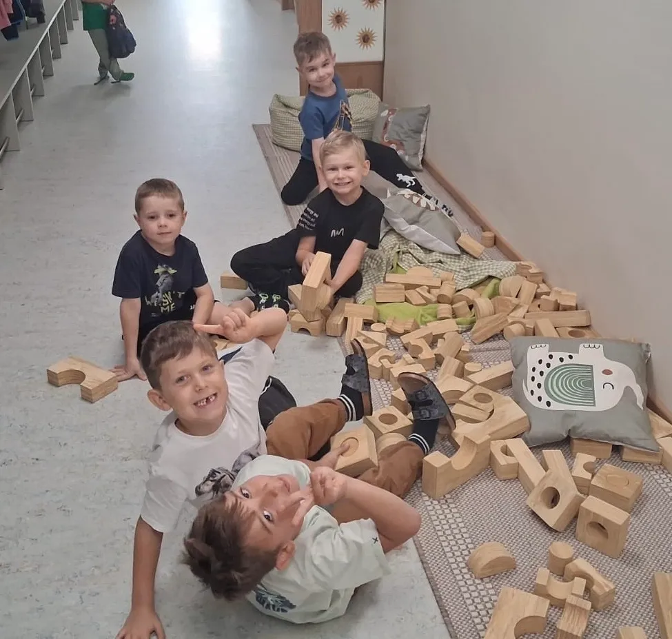 Five young boys are playing with wooden blocks in a room. One boy is lying on the floor, another is sitting, and three others are standing. A woman is standing in the background.