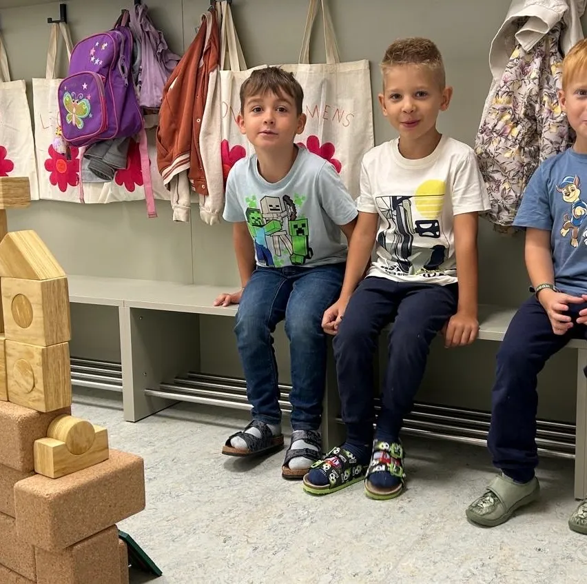 Drei junge Jungen sitzen auf einer Bank in einem Raum mit bunten Taschen, die an der Wand hängen.