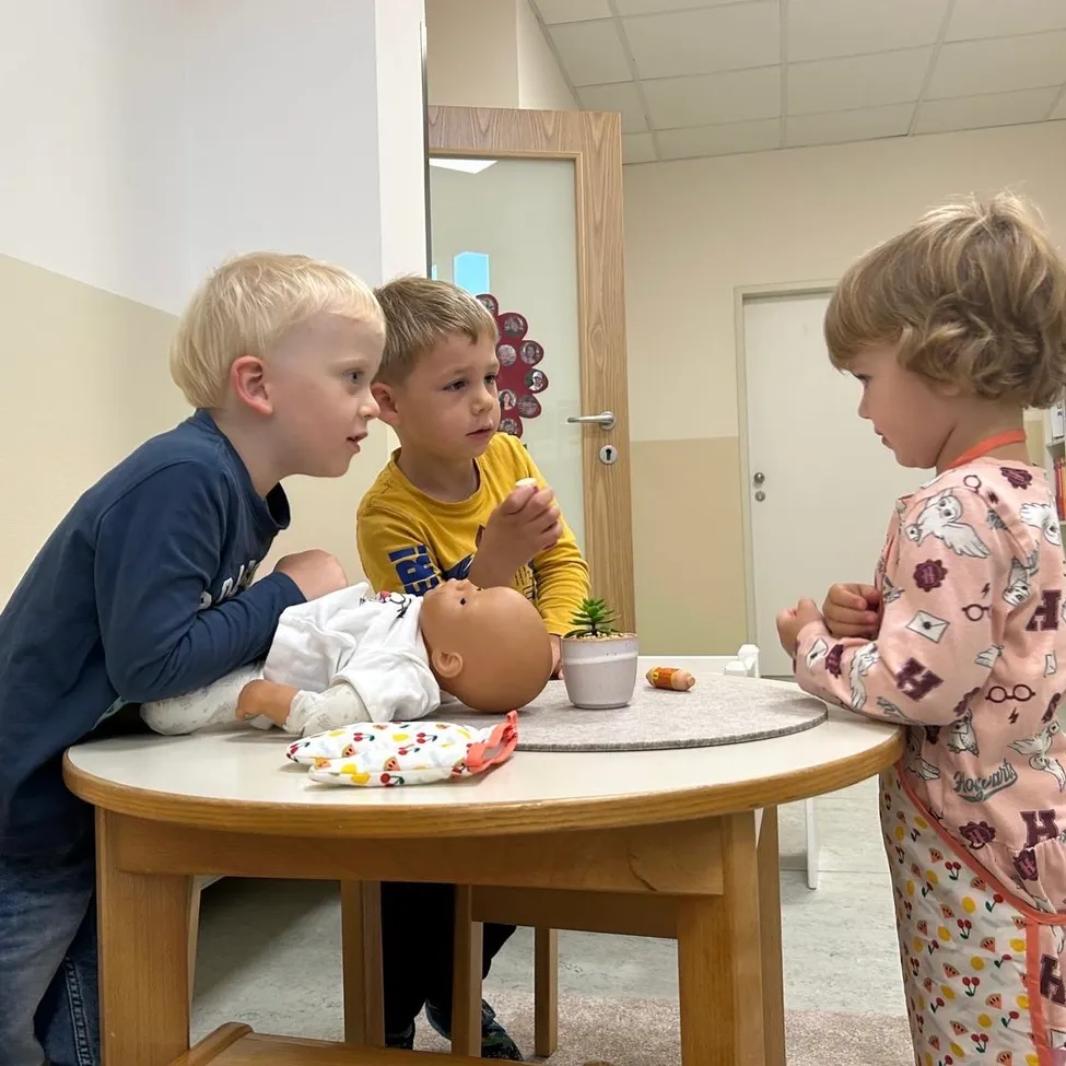 Three young children are gathered around a table, examining a baby doll. One child holds the doll while another looks on closely. A small potted plant sits on the table.