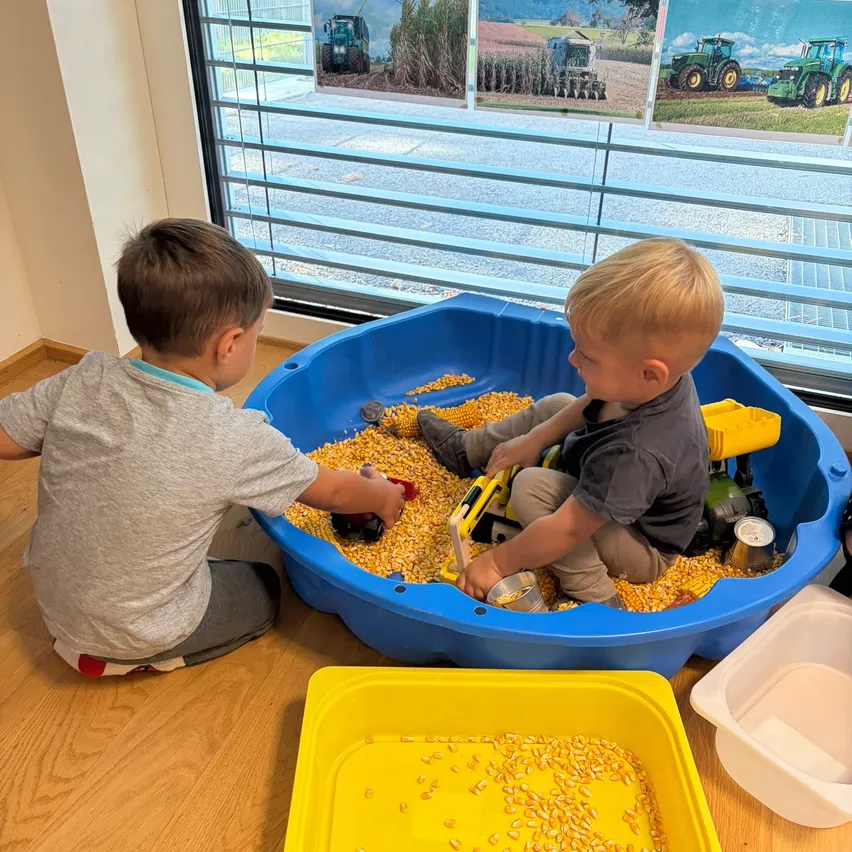 Zwei kleine Jungen spielen mit Spielzeug-Lkw in einer blauen Kiste voller Mais, sitzen auf einem Holzboden mit einem Fenster, das einen Blick auf Traktoren zeigt.