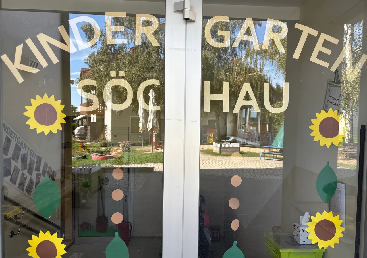 Eine Glastür mit weißem Rahmen und dekorativen Elementen. Der Text lautet 'UNTER GARTEN SÖC HAUS'. Der Hintergrund zeigt einen Garten mit einem Baum, einer Bank und einem Haus.