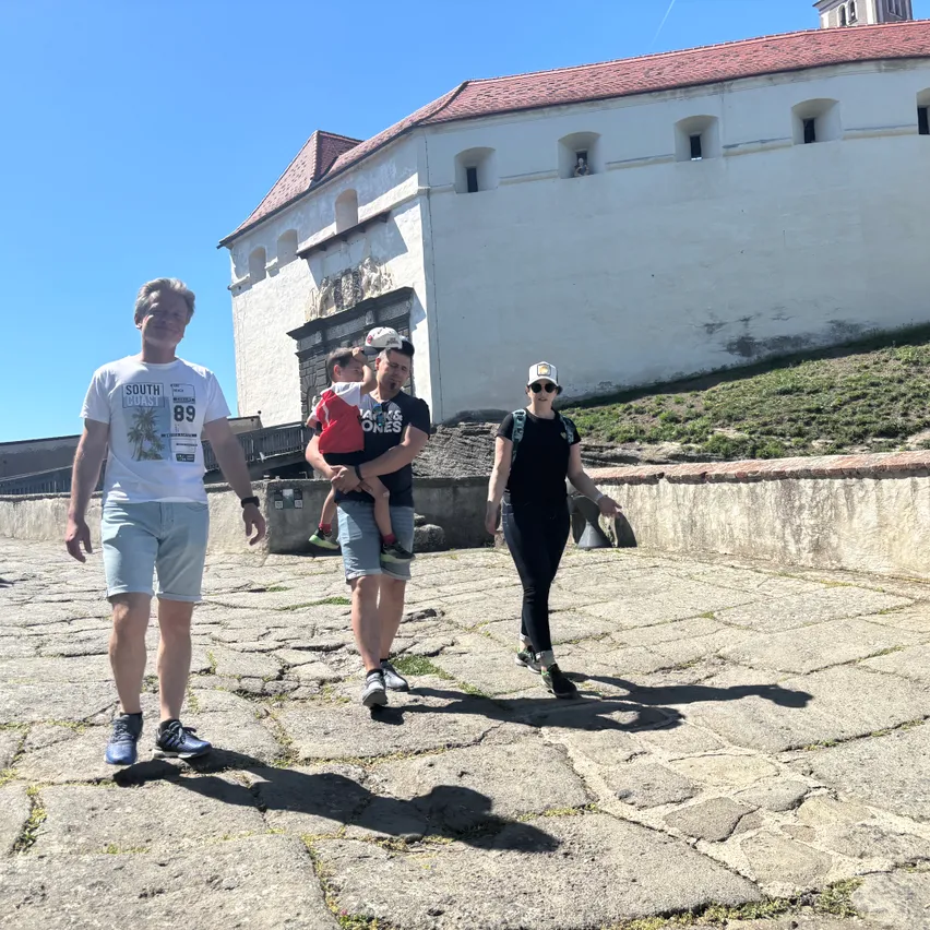 Drei Menschen laufen auf Kopfsteinpflaster in der Nähe eines Gebäudes mit rotem Dach. Ein Mann hält ein Kind, während eine Frau in Sonnenbrille neben ihnen geht.