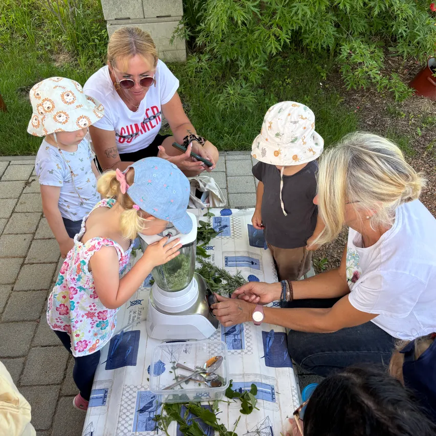 Zwei Frauen und vier Kinder stehen um einen Tisch herum, wobei zwei Kinder vor einem mit grünen Blättern gefüllten Mixer stehen. Eine Frau unterrichtet die Kinder, während die andere ein Telefon benutzt.