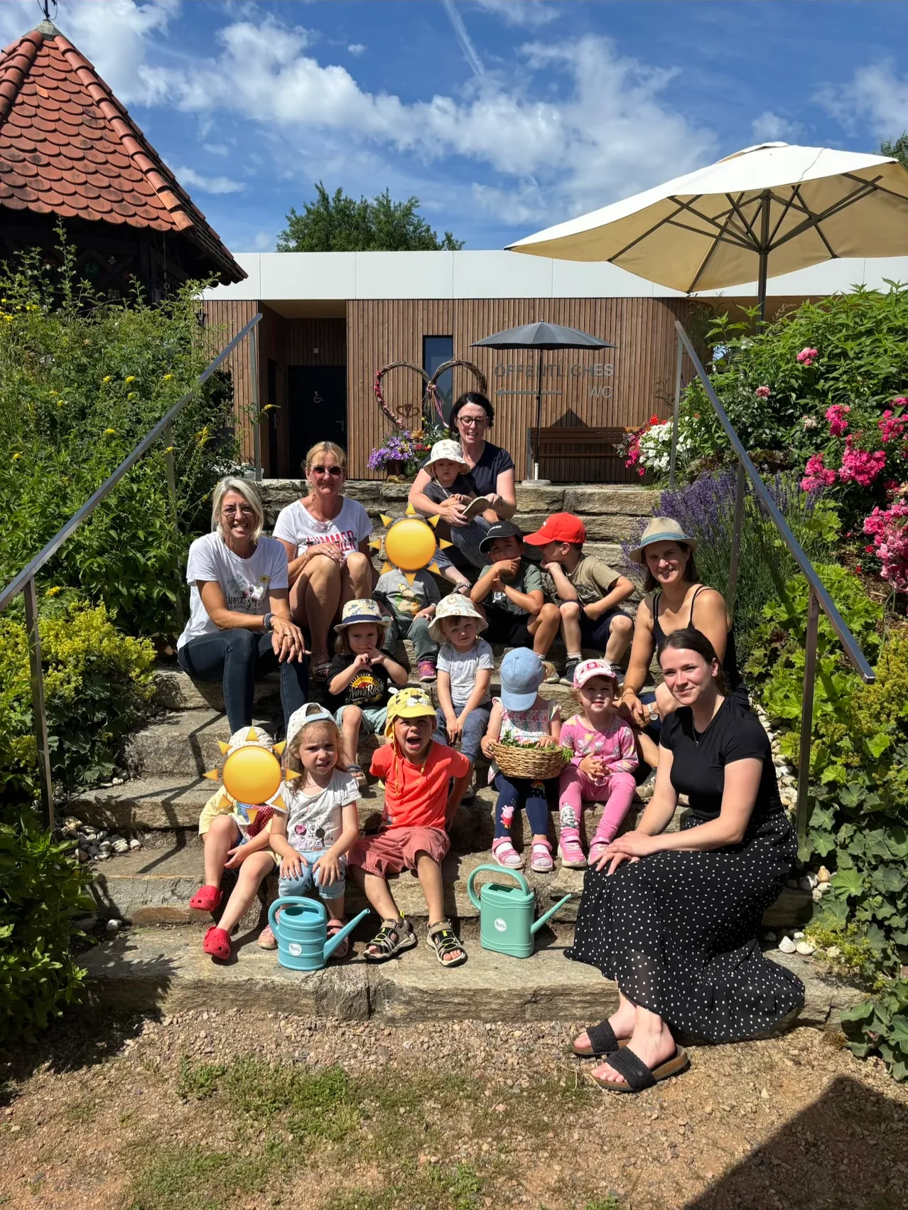 Eine Gruppe von Erwachsenen und Kindern posiert für ein Foto auf einer Gartentreppe, hält Luftballons und Gießkannen. Sie sind von Pflanzen und Blumen umgeben.