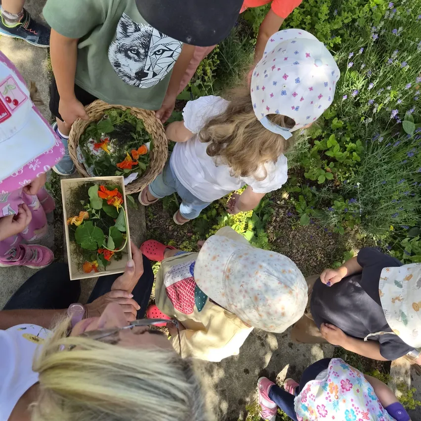 Kinder versammeln sich, um einen Korb mit Blumen und Kräutern zu untersuchen. Eine Frau mit Brille hält eine Blumenschachtel und ein Kind in der Nähe trägt einen Hut mit Blumendesigns.