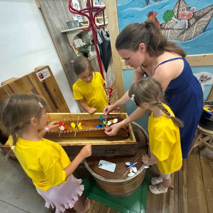 Eine Frau und drei Mädchen spielen mit bunten Spielzeugen auf einem Holztisch in einem Raum mit einem Wandbild.