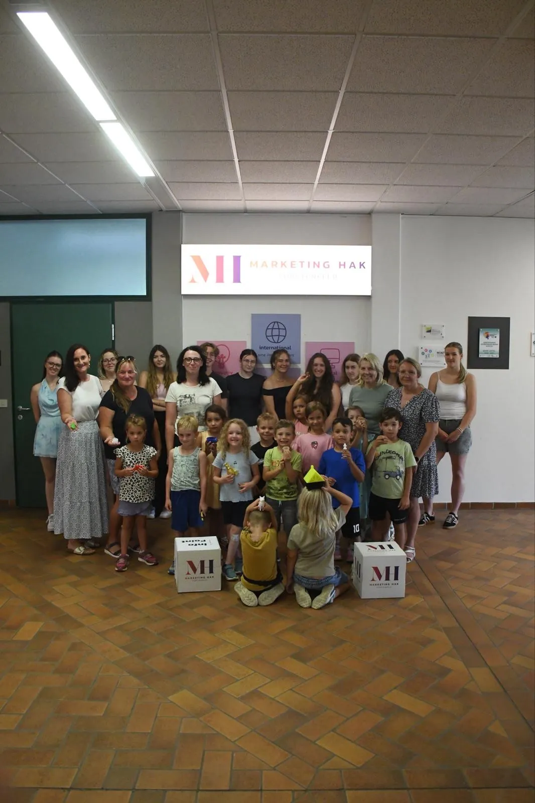Eine Gruppe von Erwachsenen und Kindern posiert in einem Raum für ein Foto. Sie stehen vor einem Schild, das Marketing Hak sagt.