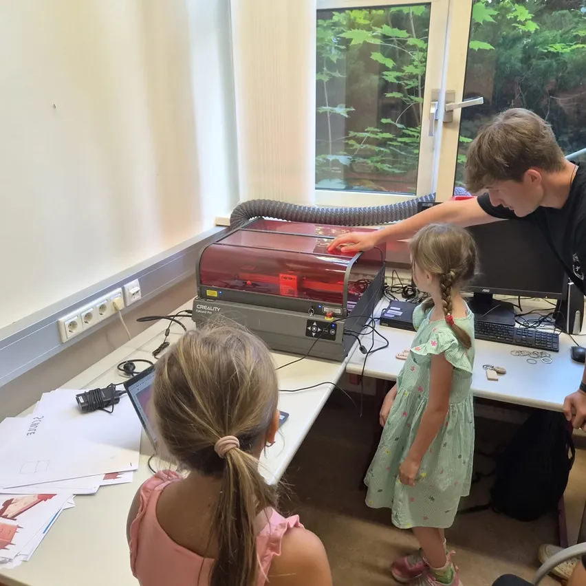Ein junger Mann bedient einen 3D-Drucker, während ein Mädchen zuschaut. Beide sind in einem Raum mit einem Schreibtisch, einem Monitor und einem Fenster. Papiere und ein Laptop sind auf dem Schreibtisch.
