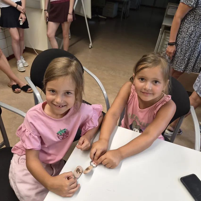 Zwei kleine Mädchen sitzen an einem Tisch und beschäftigen sich mit einer Aktivität. Sie tragen rosa Hemden und haben Schlüssel in den Händen. Der Tisch hat eine weiße Oberfläche.