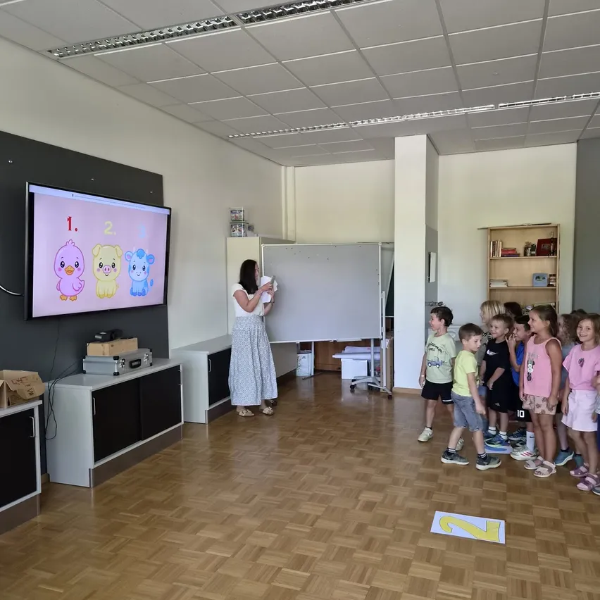 Eine Frau unterrichtet eine Gruppe von Kindern in einem Klassenzimmer. Ein Monitor zeigt Cartoon-Tiere an, und die Kinder stehen in einer Reihe.
