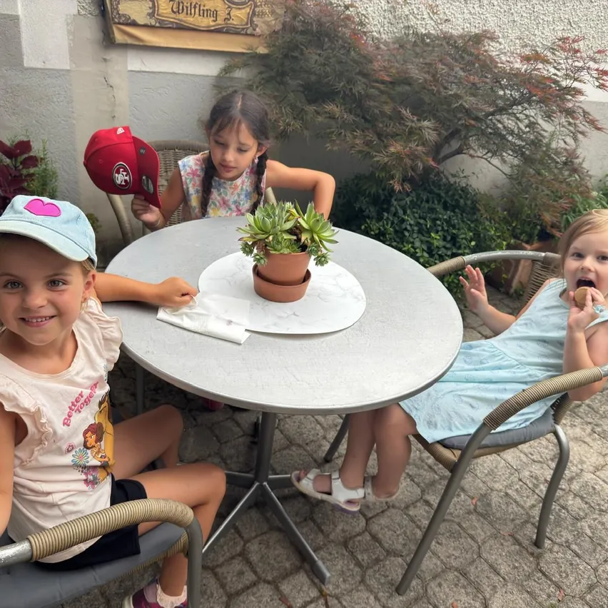 Drei junge Mädchen sitzen um einen Tisch herum, lächeln und posieren für ein Foto. Eines hält einen roten Hut. Eine Pflanze steht auf dem Tisch, und Grünpflanzen umgeben sie.