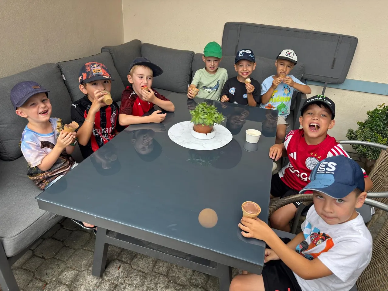 Eine Gruppe von Kindern sitzt um einen Tisch und genießt Eis und Snacks. Sie tragen alle Mützen und einige tragen Sporttrikots.