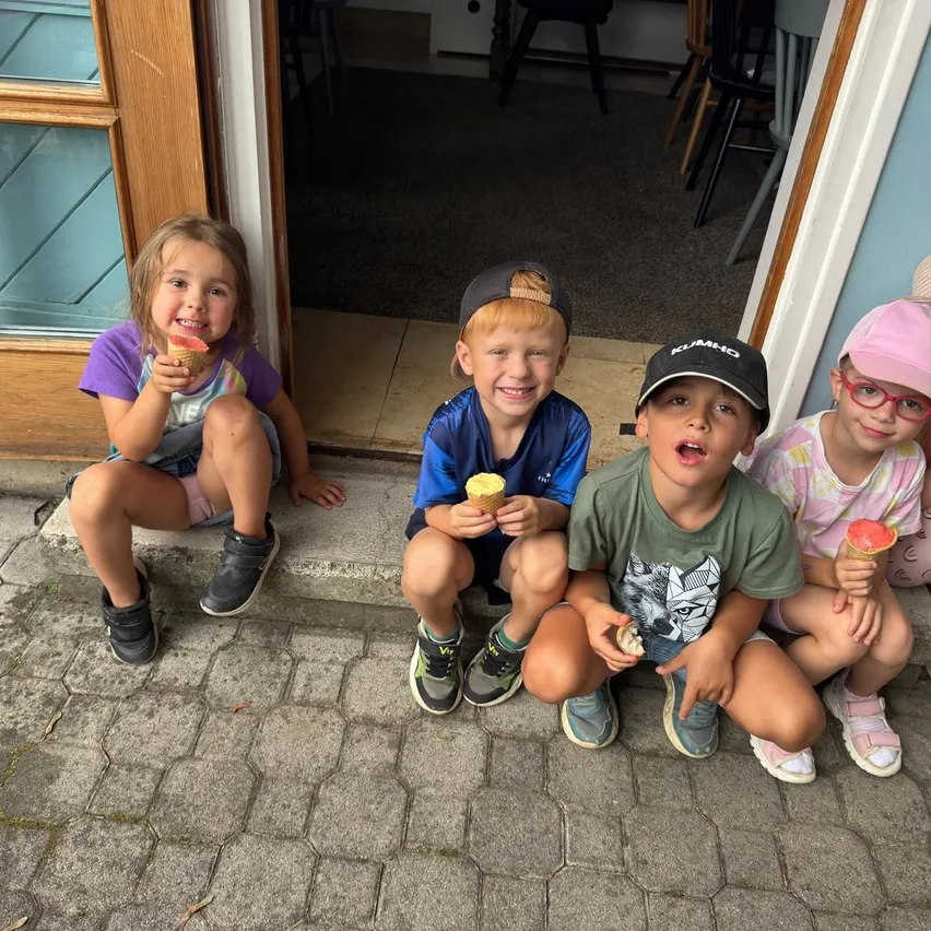 Vier Kinder sitzen auf einem Bürgersteig und halten Eisbecher in der Hand. Sie lächeln. Die Tür hinter ihnen ist offen. Der Boden ist mit Teppich ausgelegt.