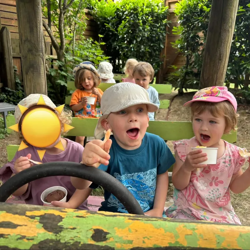Gruppe von Kindern auf grünen Bänken, die Eiscreme essen, in einem sonnendurchfluteten Garten mit einem Holzzaun und Pflanzen im Hintergrund.