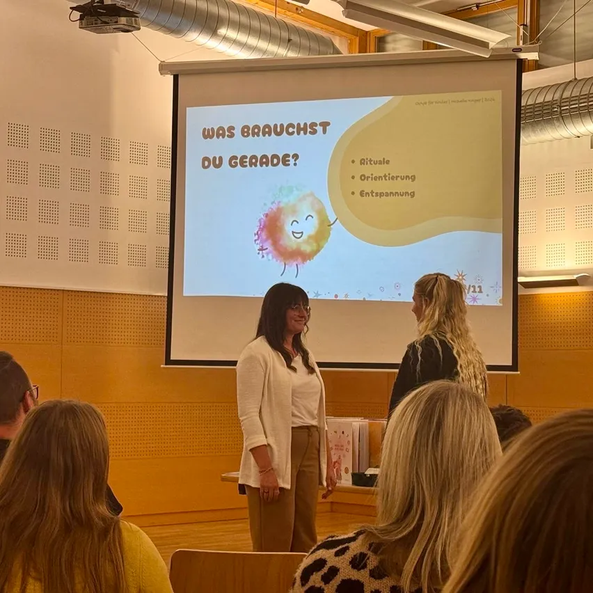 Zwei Frauen stehen vor einer Projektionsleinwand in einem Klassenzimmer mit einer Präsentation über Rituale, Orientierung und Entspannung.