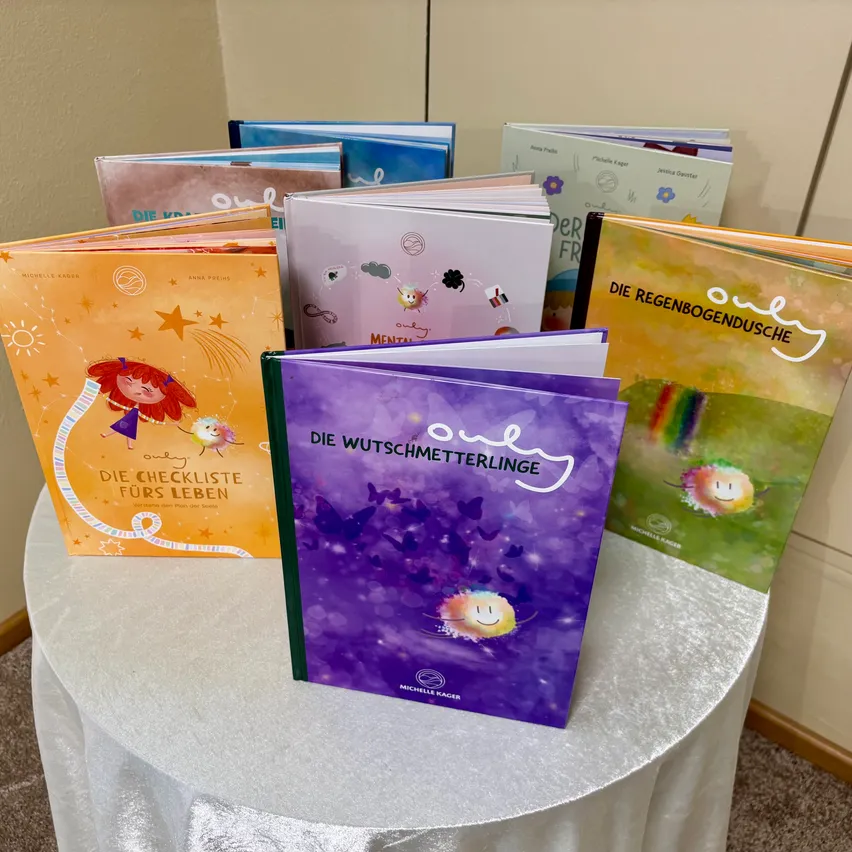 Ein Tisch, der mit einem weißen Tuch bedeckt ist, hat mehrere Bücher darauf. Die Bücher sind in einer Reihe angeordnet, und die Einbände sind farbenfroh und haben verschiedene Designs. Die Bücher haben Titel auf Deutsch und einige von ihnen haben Illustrationen.