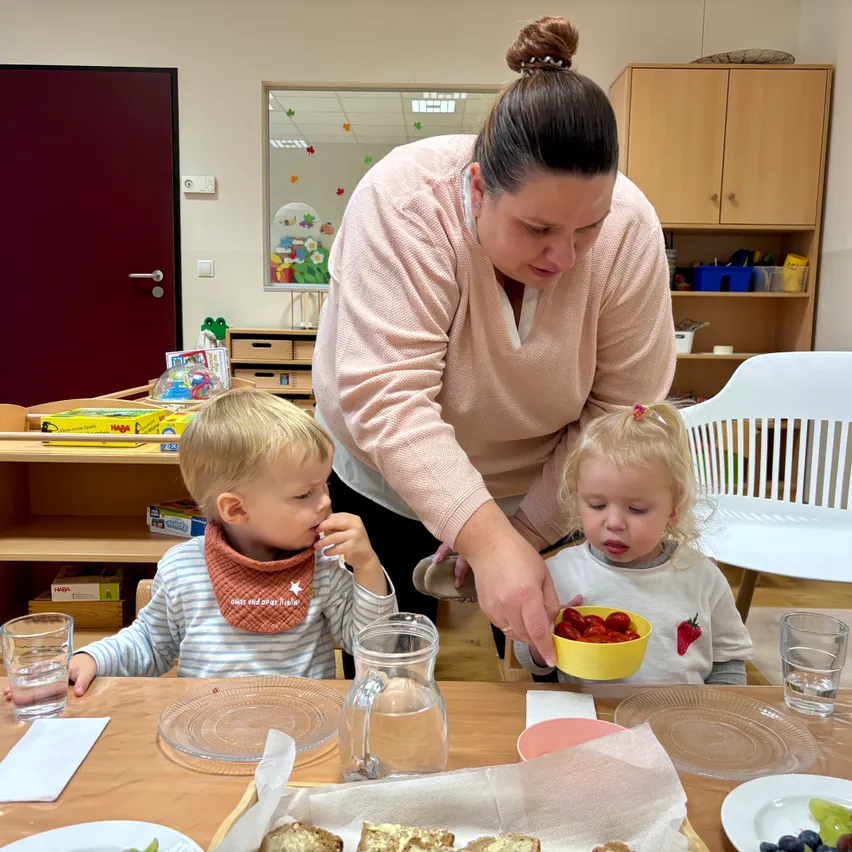 Eine Frau füttert zwei kleine Kinder am Tisch. Die Kinder essen aus Schüsseln.
