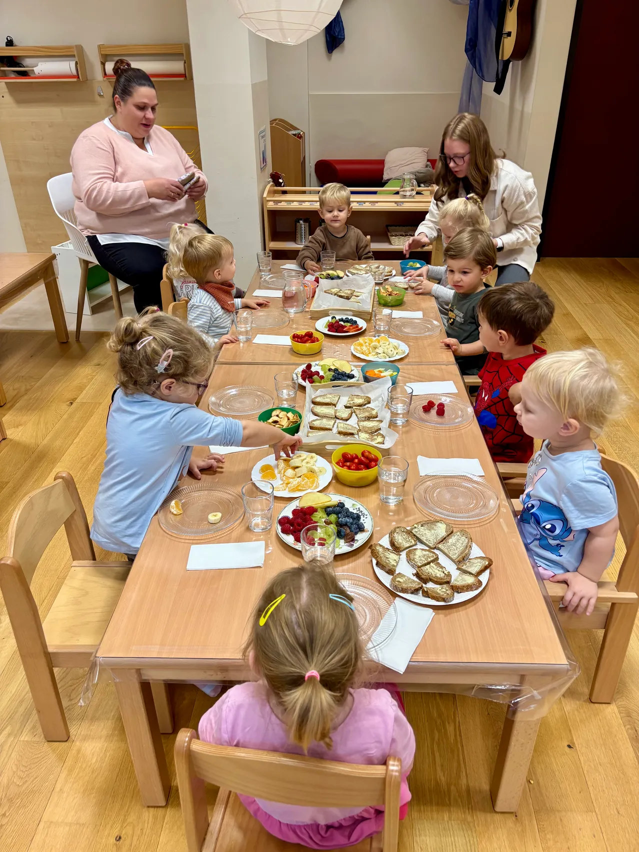 Eine Gruppe von Kindern sitzt um einen Tisch herum und isst verschiedene Lebensmittel. Eine Frau steht hinter ihnen und eine andere Frau sitzt in der Nähe.