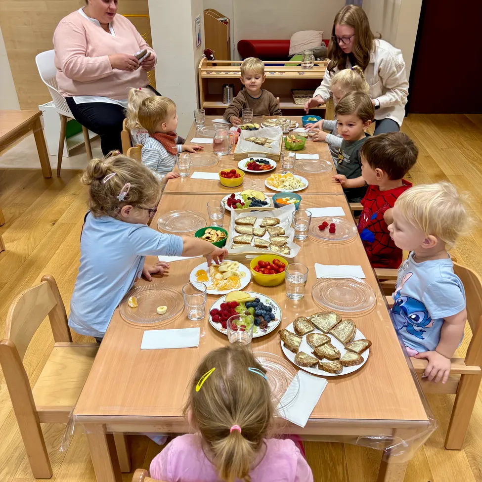 A group of children are sitting around a table, eating various foods. A woman is standing behind them, and another woman is sitting nearby.