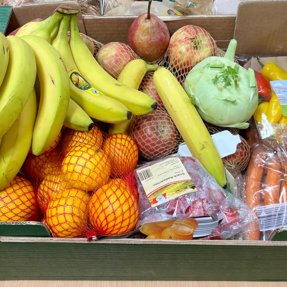 A green box filled with various fruits and vegetables, including bananas, apples, oranges, carrots, and peppers.