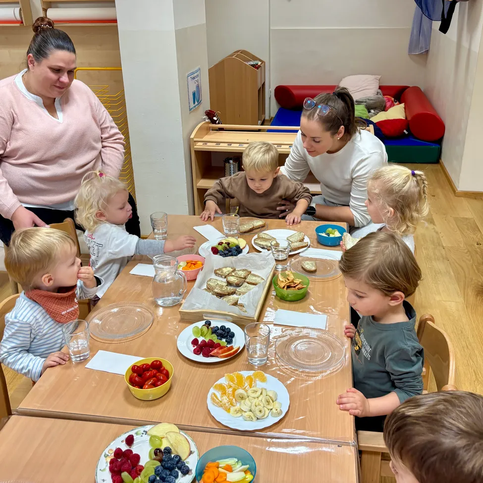 A group of children and two adults are sitting around a table in a room, eating food.