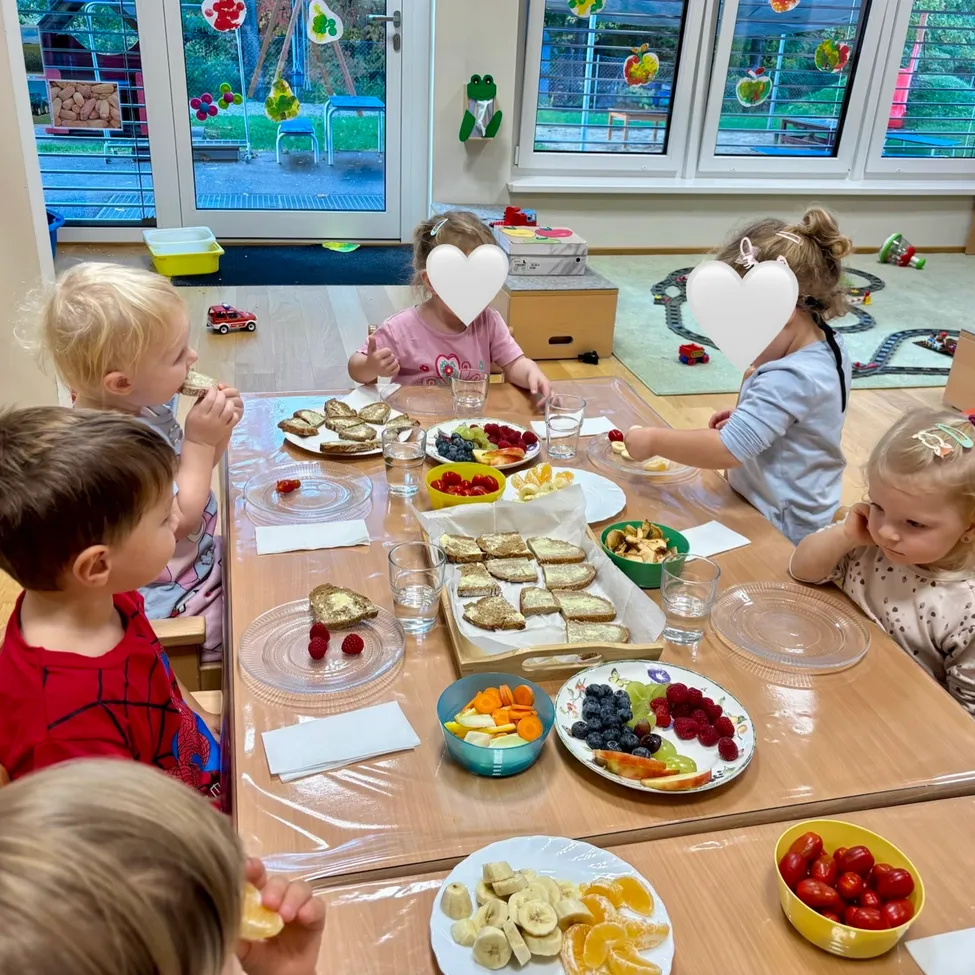 Five children are sitting around a table in a room, eating food. The table is covered with plastic and has plates of fruits, sandwiches, and drinks.
