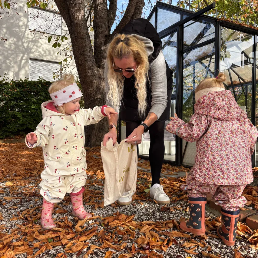 A woman and two young children are standing on a gravel path. The woman is holding a white bag, and the children are wearing pink rain suits.