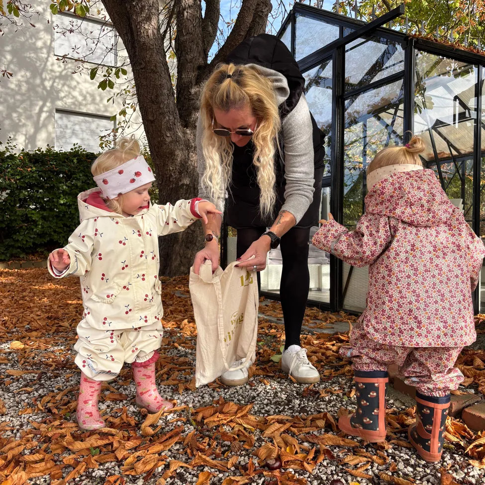 Eine Frau und zwei kleine Kinder stehen auf einem Kiesweg. Die Frau hält eine weiße Tasche und die Kinder tragen rosa Regenanzüge.