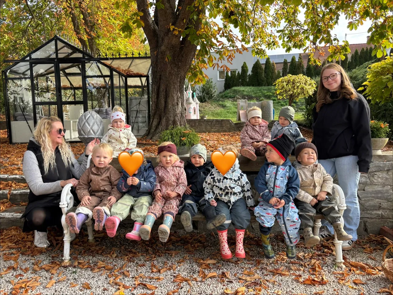 A group of children and two women are sitting on a bench in a garden. The children are holding orange heart-shaped objects.