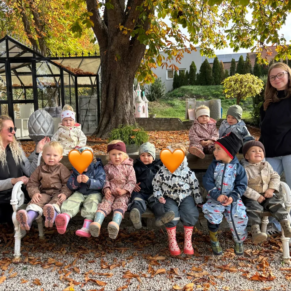 Eine Gruppe von Kindern und zwei Frauen sitzen auf einer Bank im Garten. Die Kinder halten orangefarbene herzförmige Objekte.