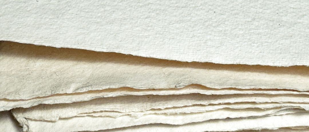 Ein Stapel gefalteter, cremefarbener Papierblätter. Die Textur ist rau und uneben, mit sichtbaren Falten und Knicken. Die Blätter scheinen handgemacht zu sein.