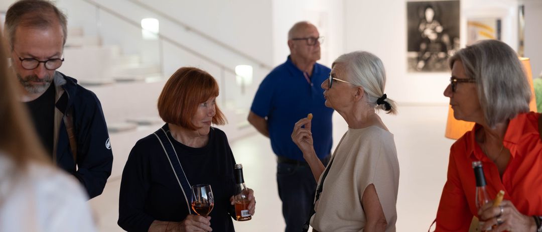 Drei Menschen stehen in einer Galerie, zwei Frauen vorn halten Wein und Snacks und ein Mann steht hinter ihnen.