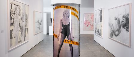 Eine Museumsgalerie mit weißen Wänden, die ein hohes zylindrisches Objekt mit einem Bild von Marilyn Monroe und Text darum herum zeigt.