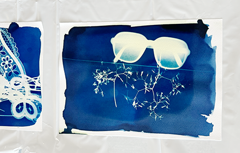Zwei Cyanotypie-Drucke auf weißem Papier. Links zeigt einer ein Spitzendesign, der Rechte eine Brille mit Ästen. Beide sind blau mit weißen Designs.
