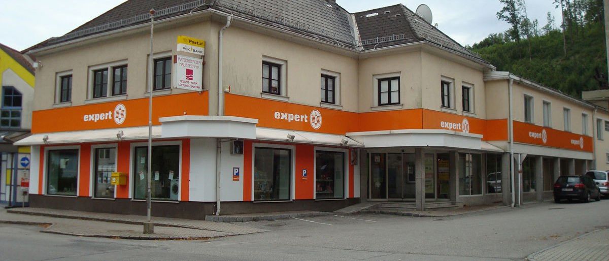 Außenansicht eines zweistöckigen Gebäudes in Orange und Weiß. Es hat das Wort 'expert' in weißer Schrift auf der orangefarbenen Wand. Eine Satellitenschüssel ist auf dem Dach. Ein Schild hängt über dem Eingang.