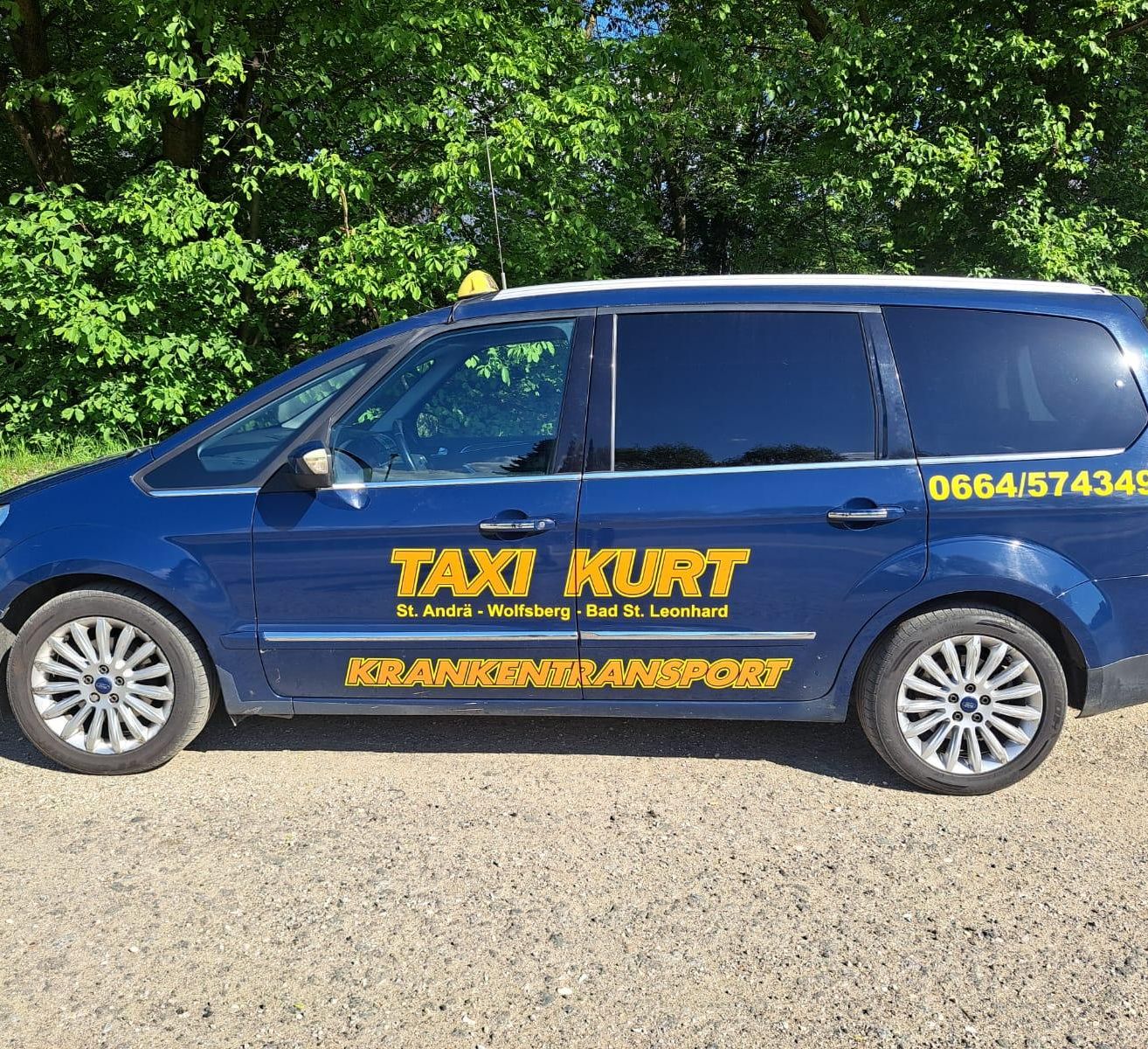 Ein blauer Ford S-MAX Van mit Taxi-Service-Werbung steht auf einer Schotterstraße mit Bäumen im Hintergrund. Die Nummer 0664/574 ist auf der Seite sichtbar.