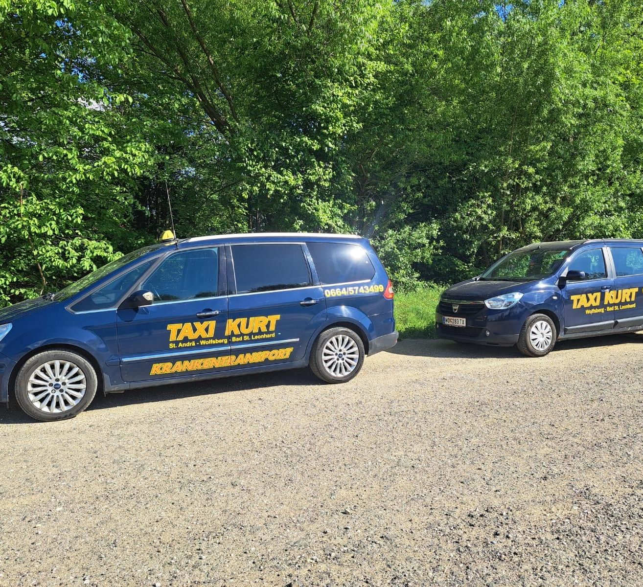 Zwei blaue Taxis auf einem Kiesweg mit Bäumen im Hintergrund geparkt. Ein Taxi hat die Aufschrift 'TAXI KURT' an der Seite.